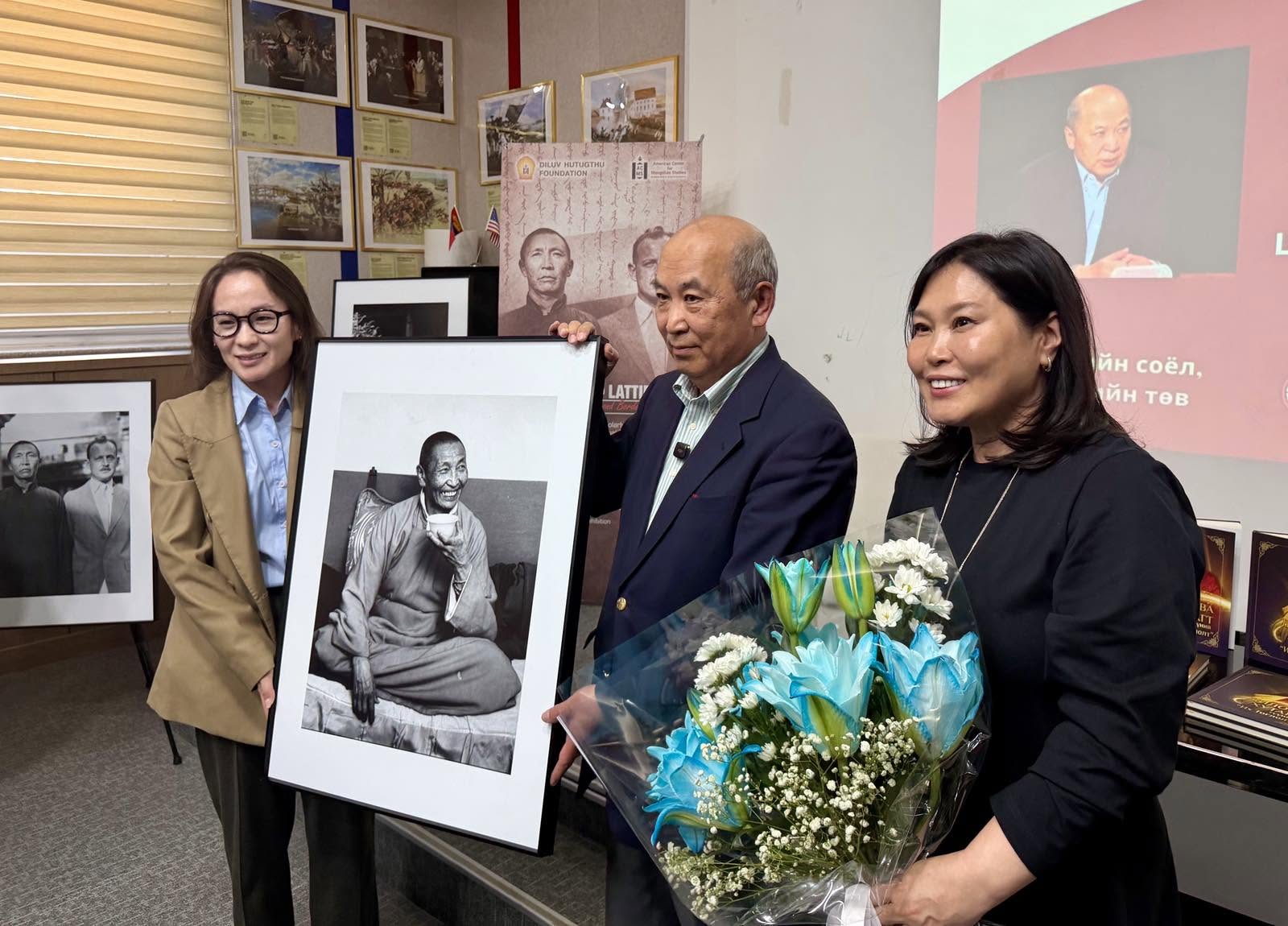 A photo of an older Mongolian man with gray hair holding a large printed and framed photograph with two Mongolian women standing beside him.