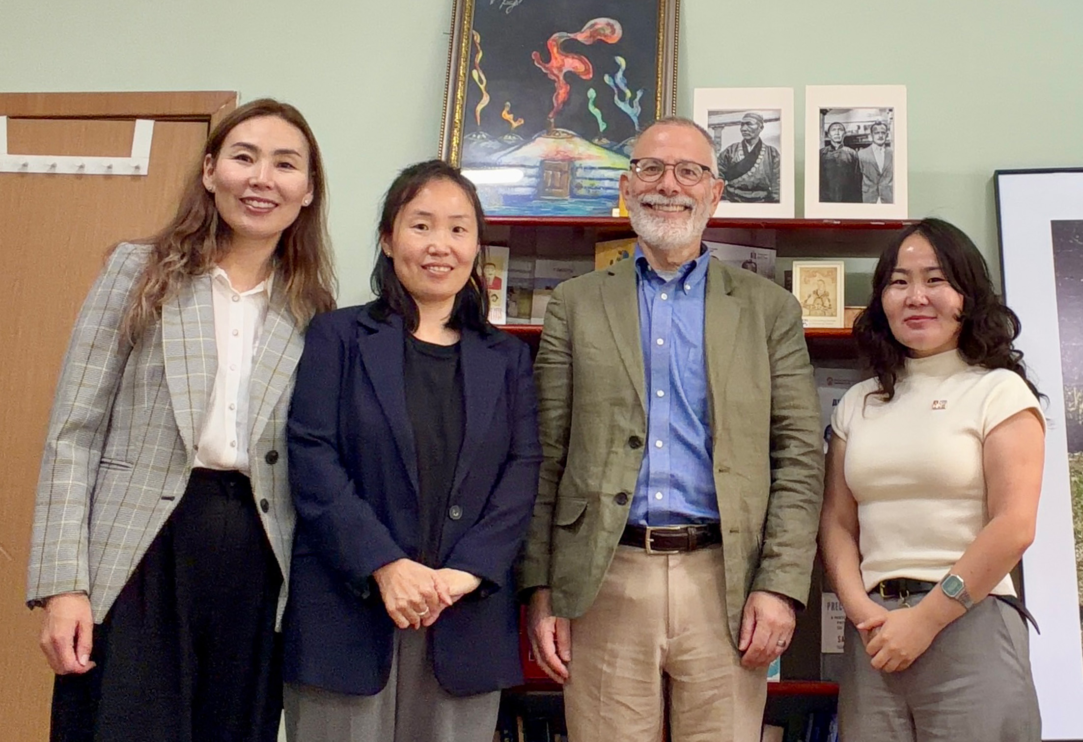 Two Mongolian women in their late thirties/early forties and one Mongolian woman in her early thirties stand on either side of an older American man. All four stand and smile looking at the camera.