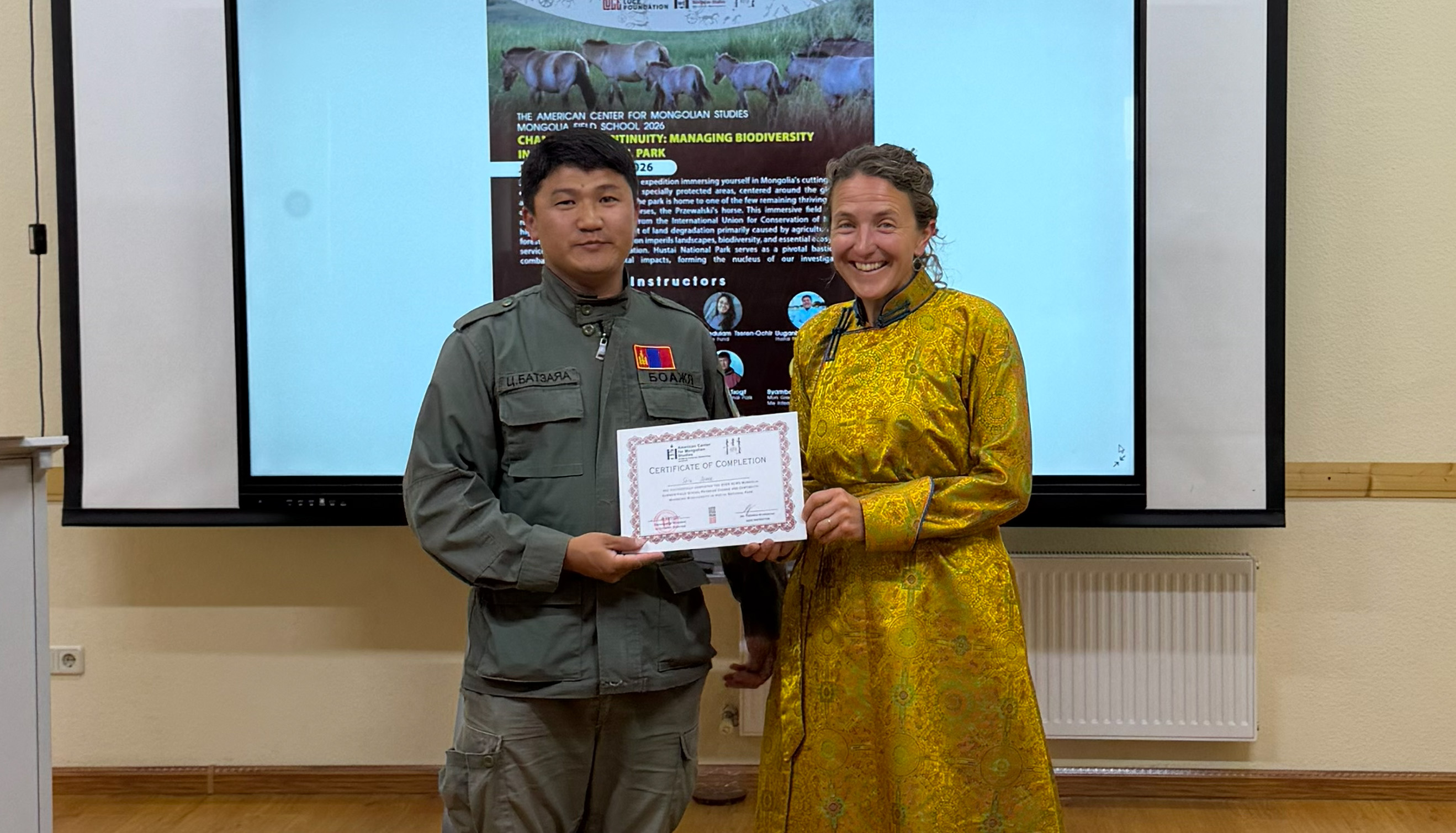 A Mongolian man in a park ranger uniform and a white woman wearing a traditional Mongolian dress stand in front of a projector screen holding a certificate.