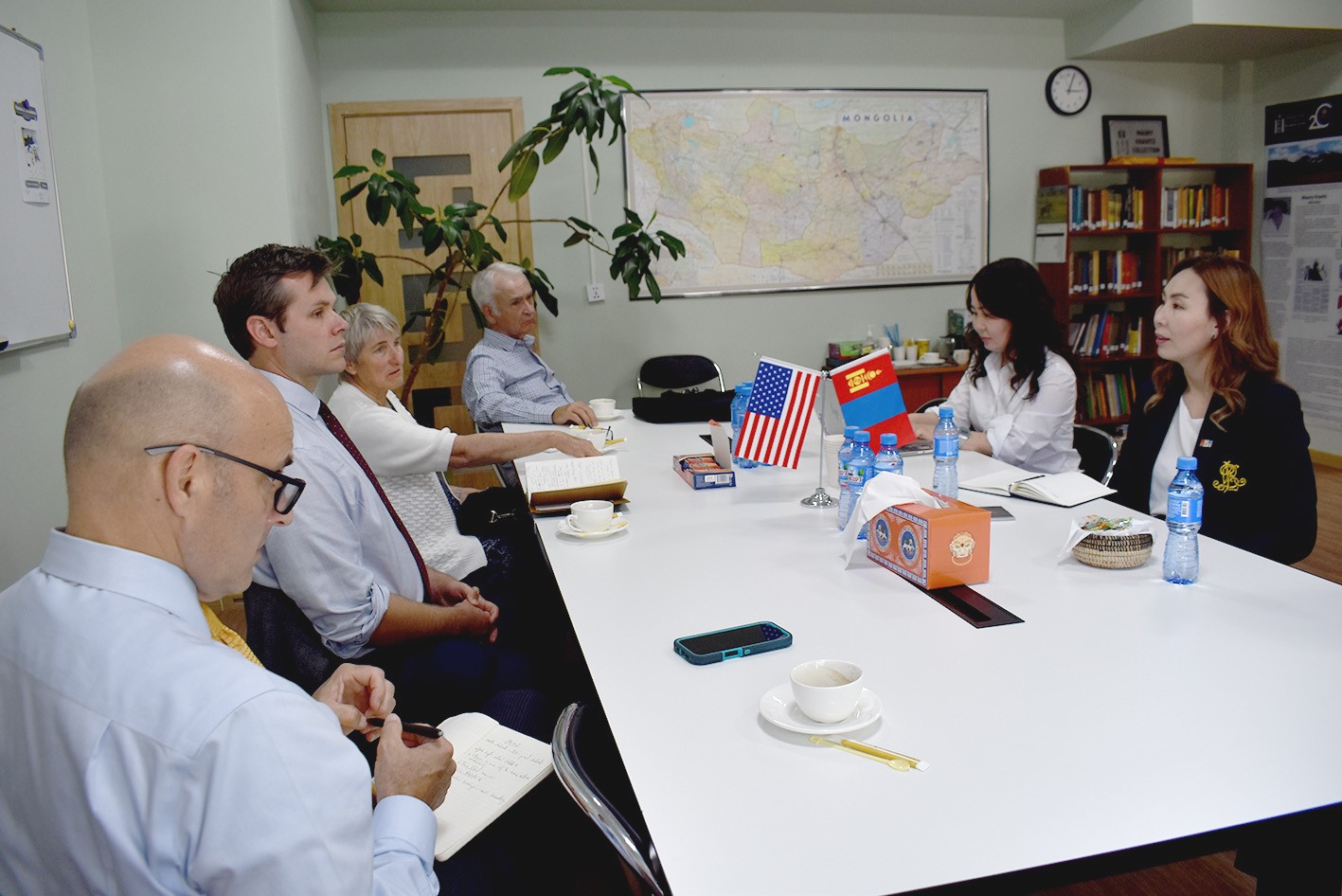 A group of American and Mongolian professionals and academics sit and discuss around a table with small American and Mongolian flags on it.