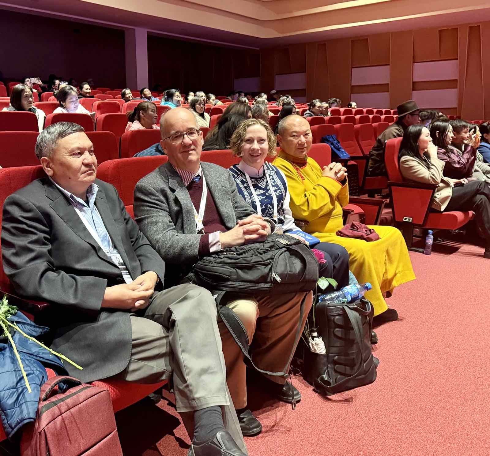 A photo of four people - three men and one woman - sitting in seats in a theater and smiling at the camera.