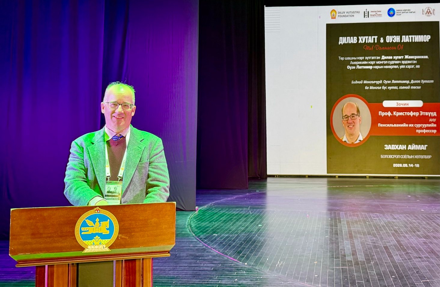 An older white man wearing a suit standing behind a podium smiling at the camera.