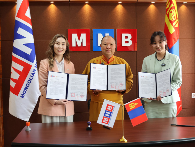 Three people stand in an office with a sign saying “MNB“ holding signed documents up for the camera.