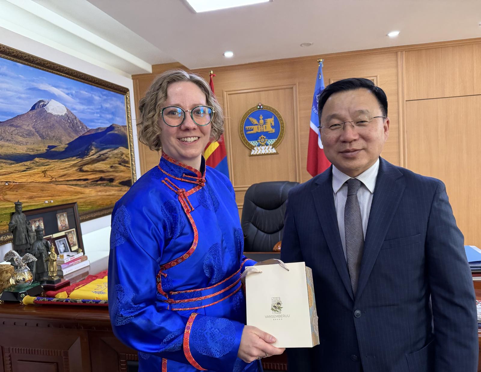 A white woman in a blue Mongolian dress holds a bag and stands next to a Mongolian man in an office. 