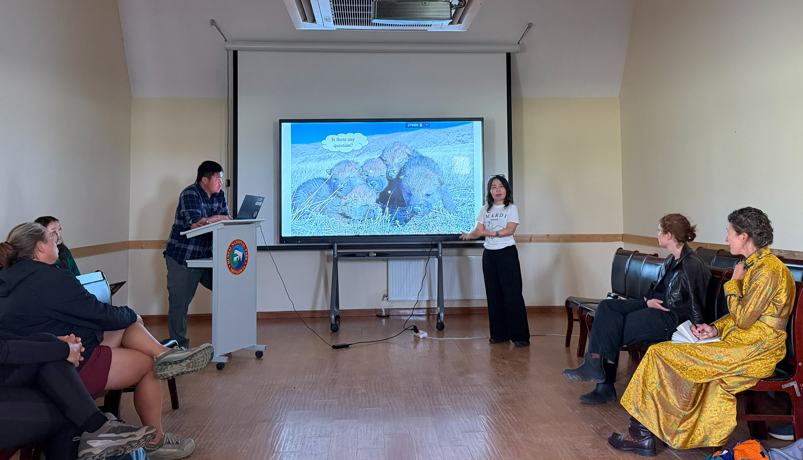 A group of five Americans sit in a classroom and listen attentively to a Mongolian woman in her thirties who is standing in front of a projector screen giving a presentation.
