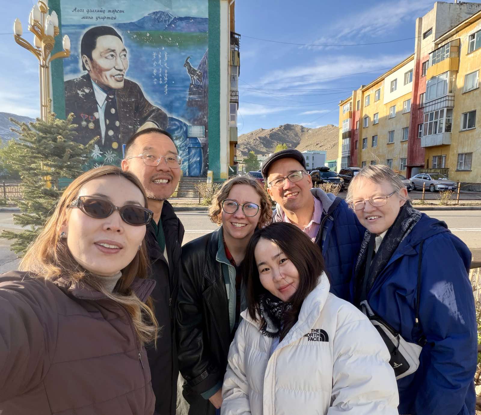 A group of four women and two men posing outside in front of a mural smiling at the camera.