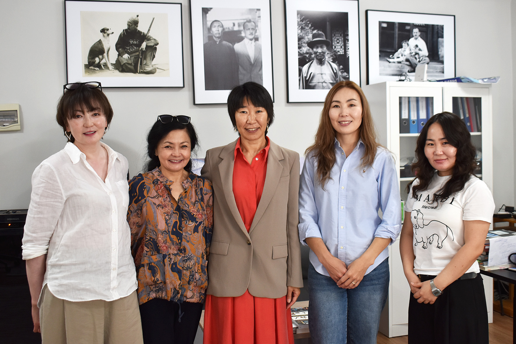 Four Mongolian women in business-casual clothing stand on either side of a Buryat woman in an orange dress and tan blazer. All five are smiling and posing together for the camera.