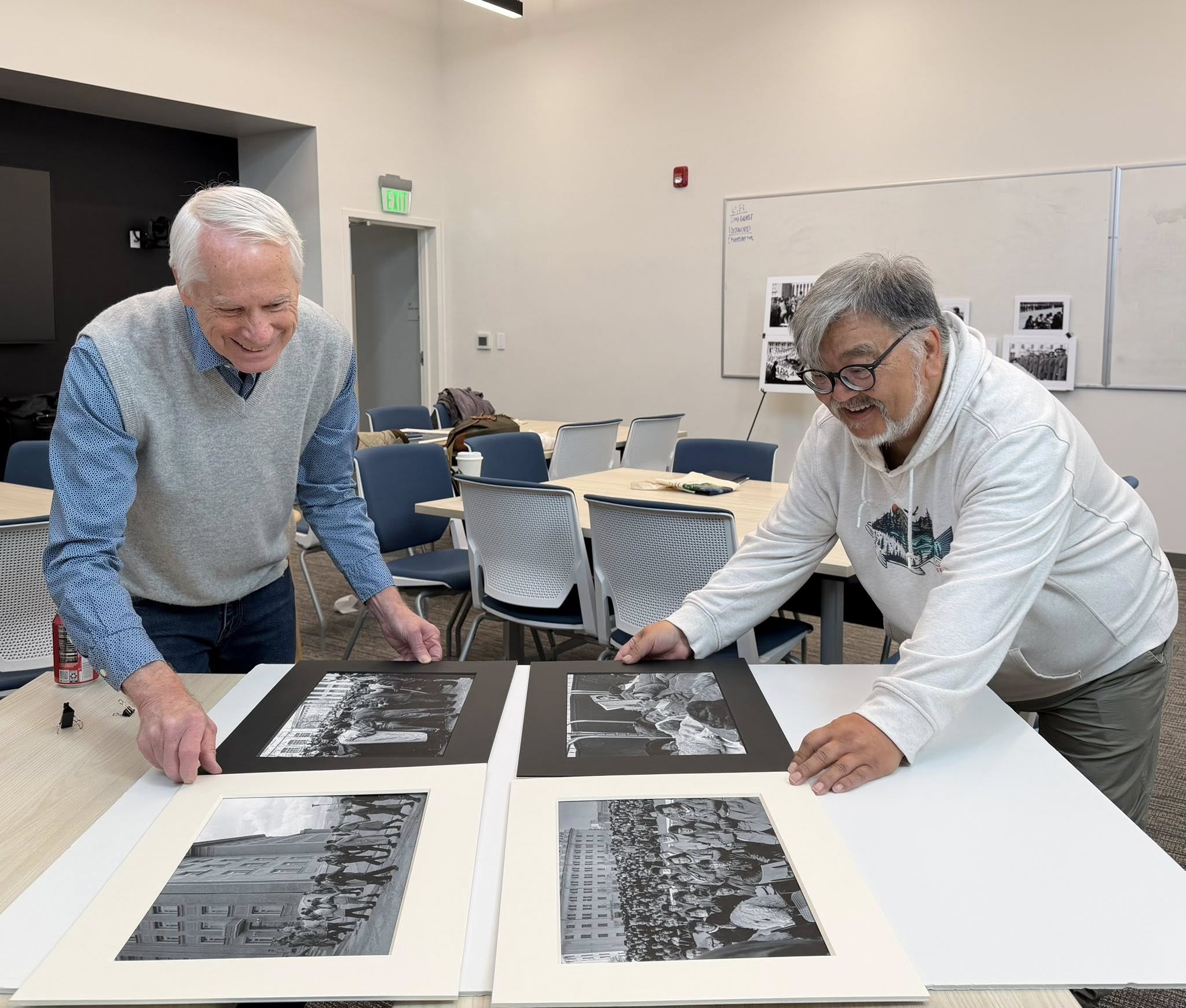 Two older men lean over a table, one showing the other photographs that he has taken.