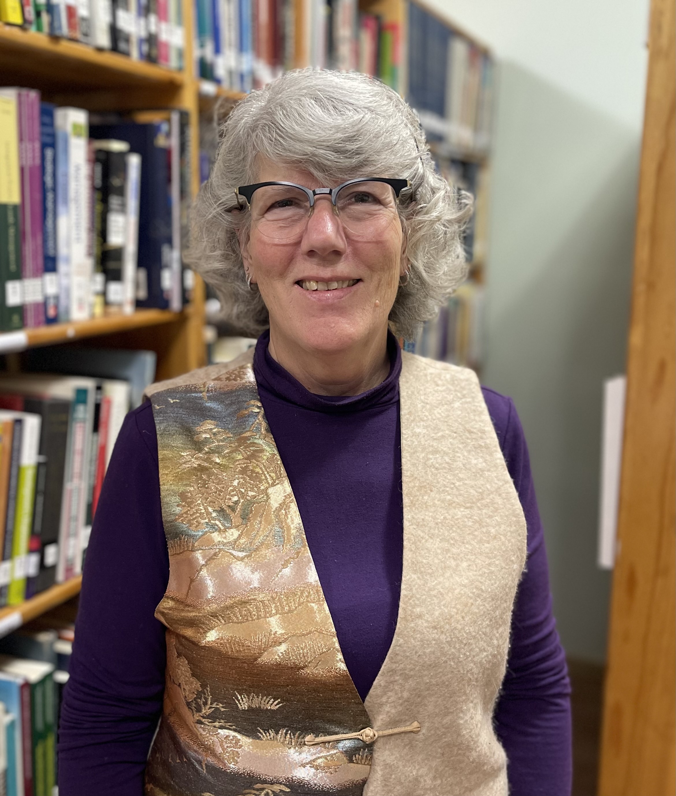 A photo of a woman with gray hair standing in front of a bookshelf and smiling at the camera.