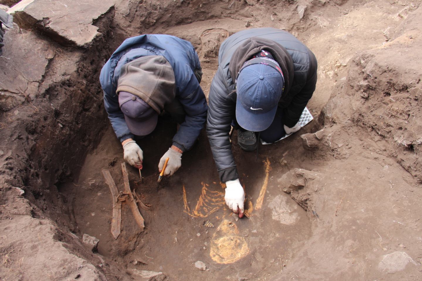 A photograph of two archaeologists kneeling in an excavated site and carefully uncovering remains.