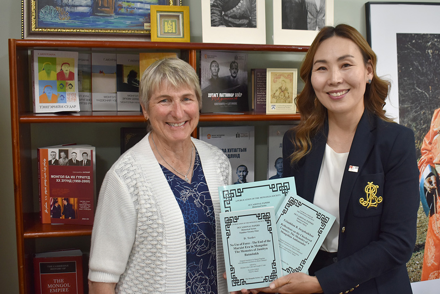 Ambassador Pamela Slutz, an older white woman, stands with ACMS In-Country Director Davaasuren Myagmar, a Mongolian woman in her thirties. They are standing together holding books and smiling for the camera.