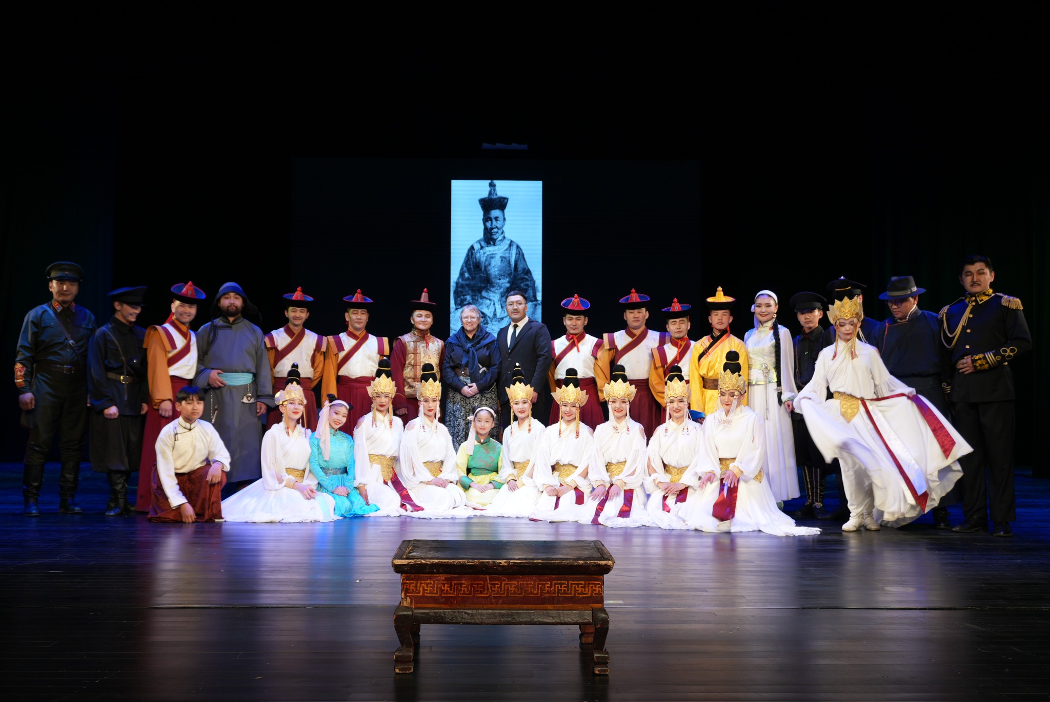 A group of performers in traditional Mongolian clothing standing onstage, posing and smiling for the camera.