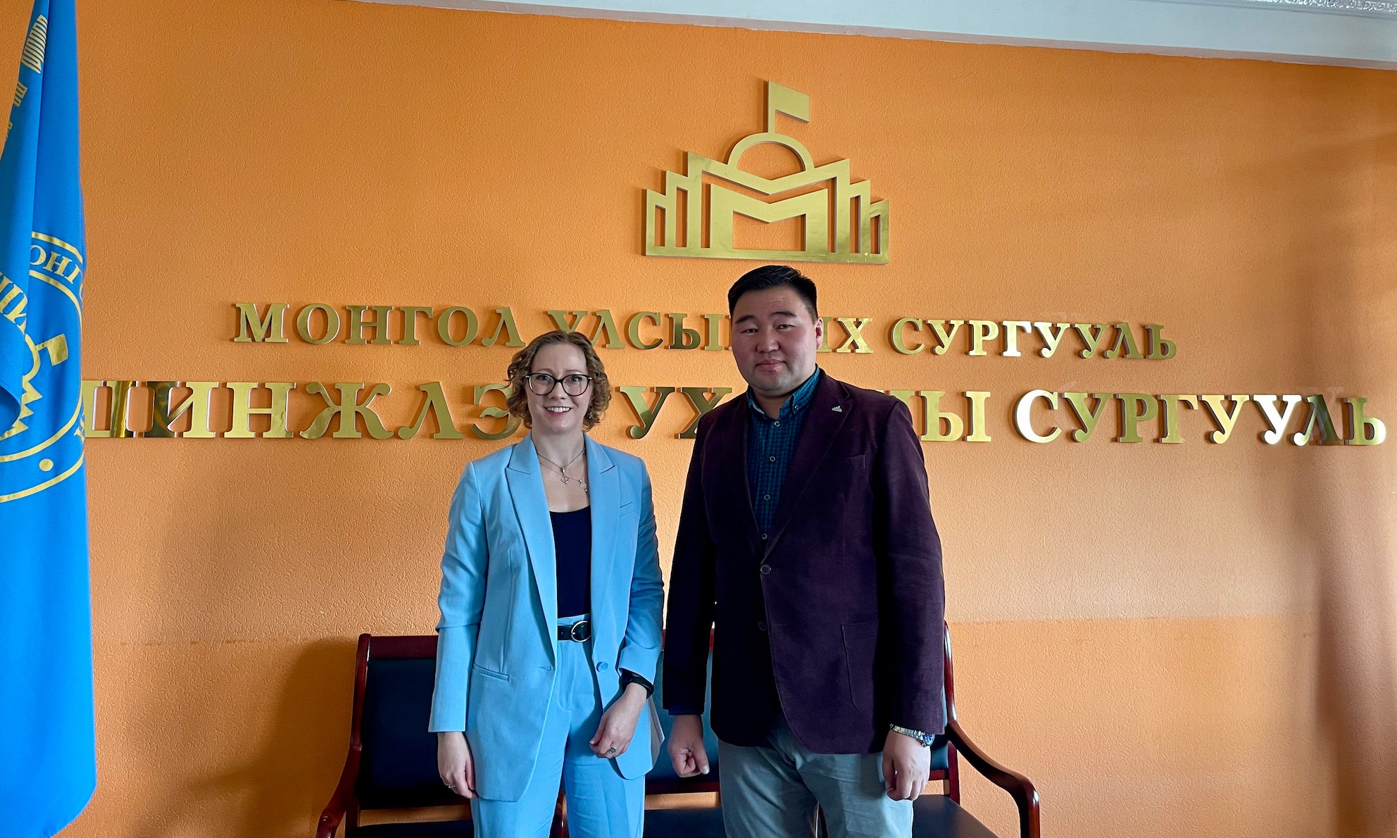 Maggie Lindrooth (left) and Associate Professor Erdene-Ochir Tumen-Ochir (right) stand in front of a wall with the National University of Mongolia logo.