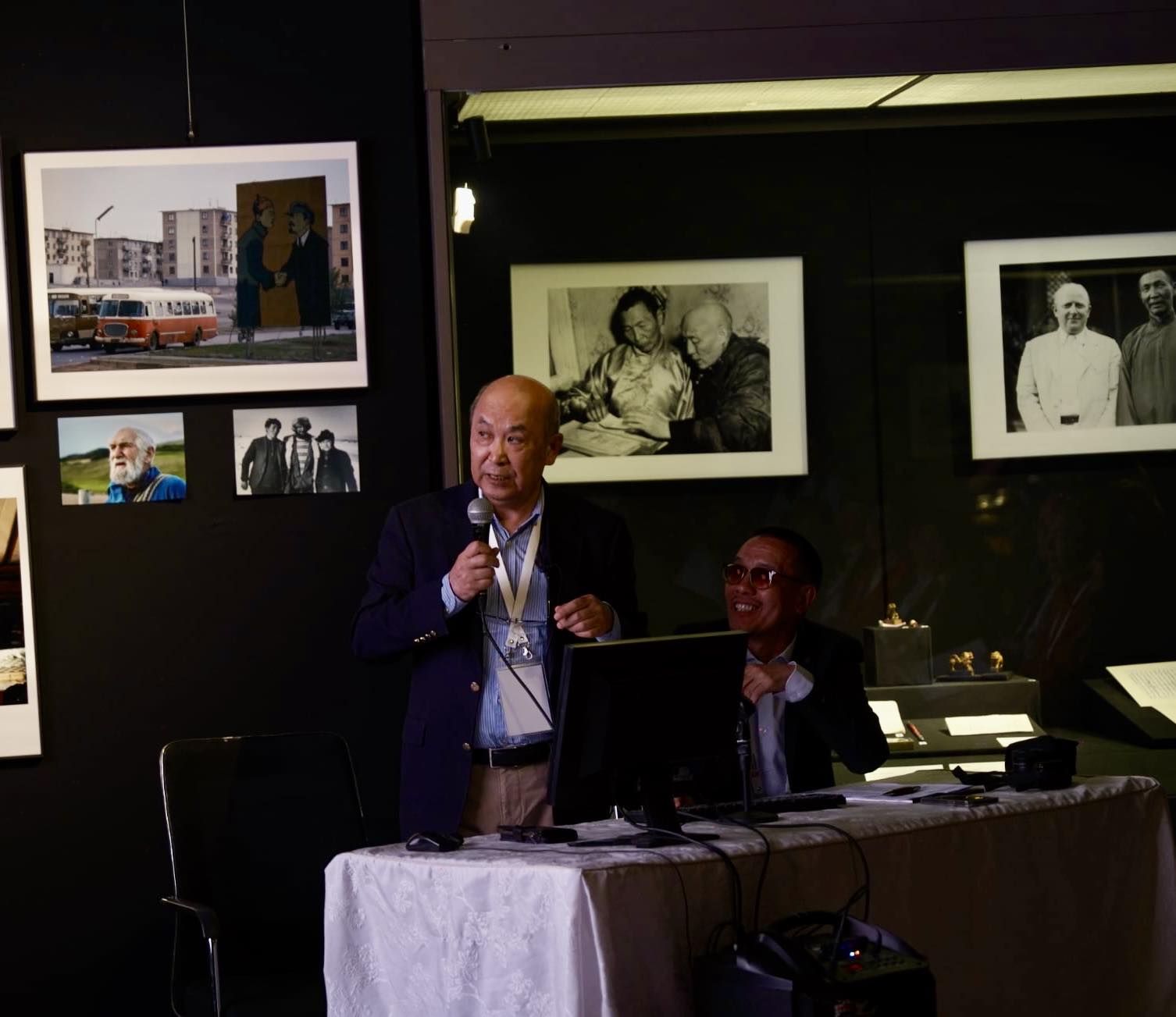 An older, bald man standing behind a desk and giving a presentation.