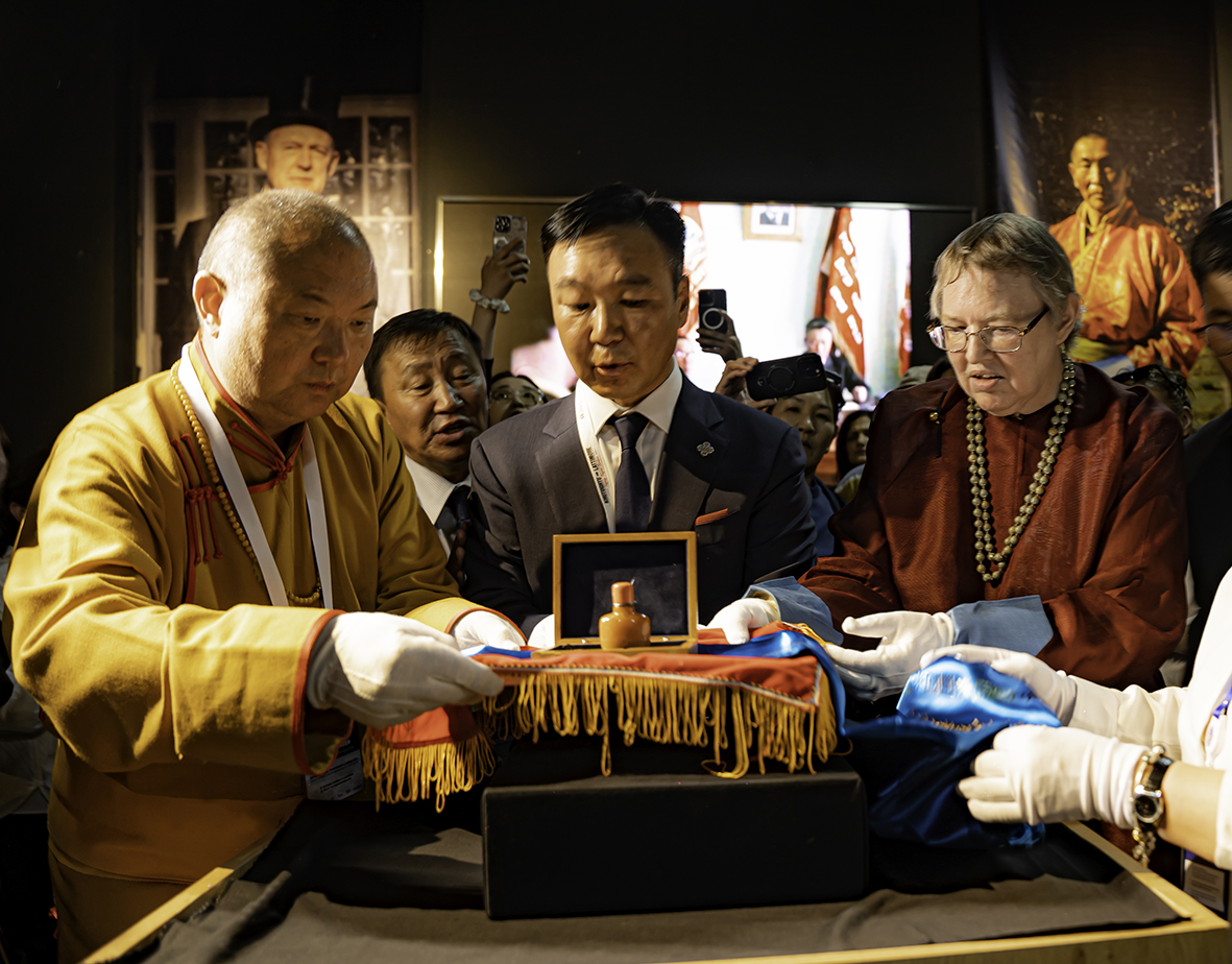 A closeup photo of three people - two in traditional Mongolian clothing and one wearing a Western suit - presenting a Mongolian snuff bottle to a museum.