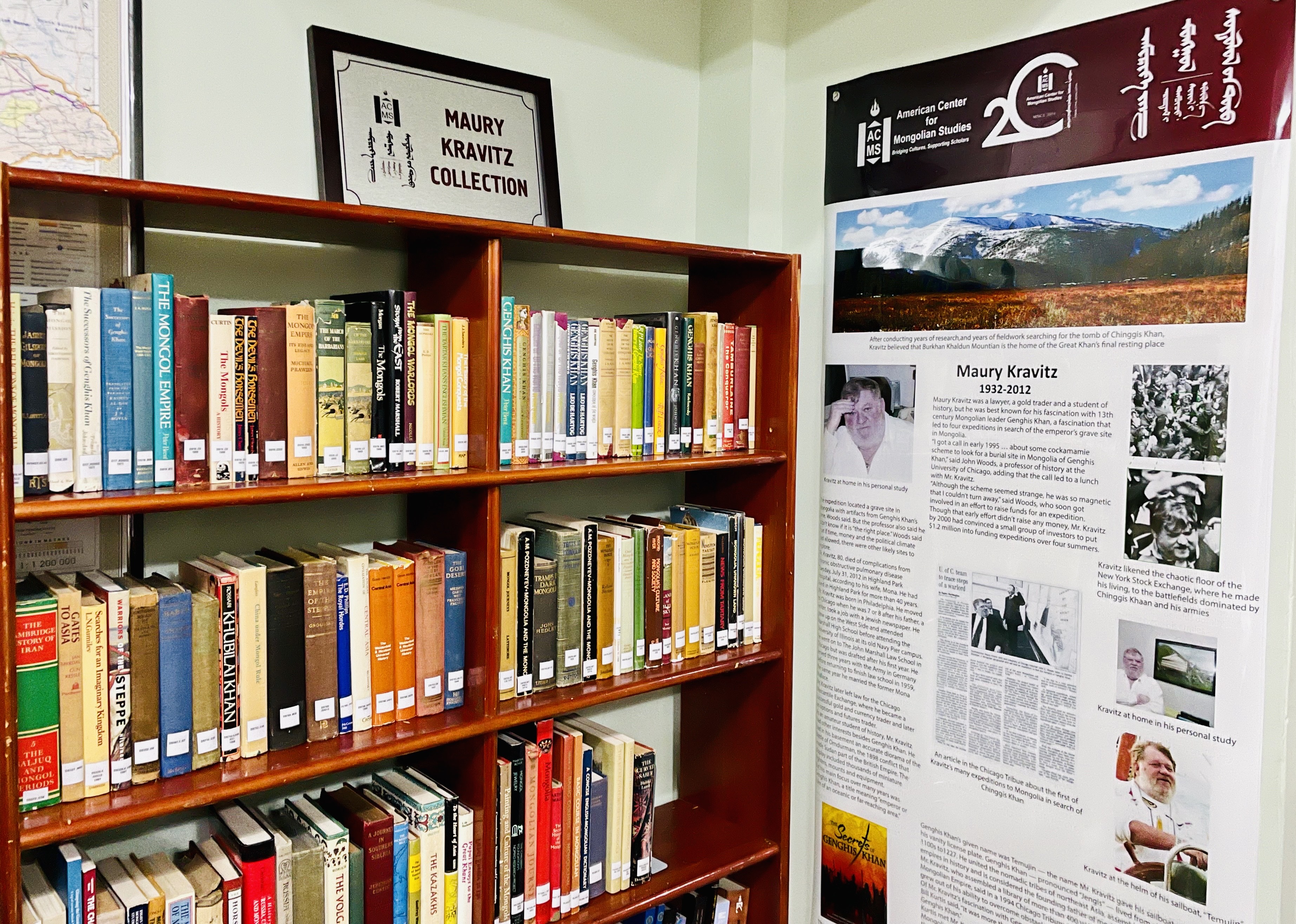 A shelf full of books with a plaque hung above it stating “Maury Kravitz Collection.“ Next to the shelf is an informational poster about Maury Kravitz.