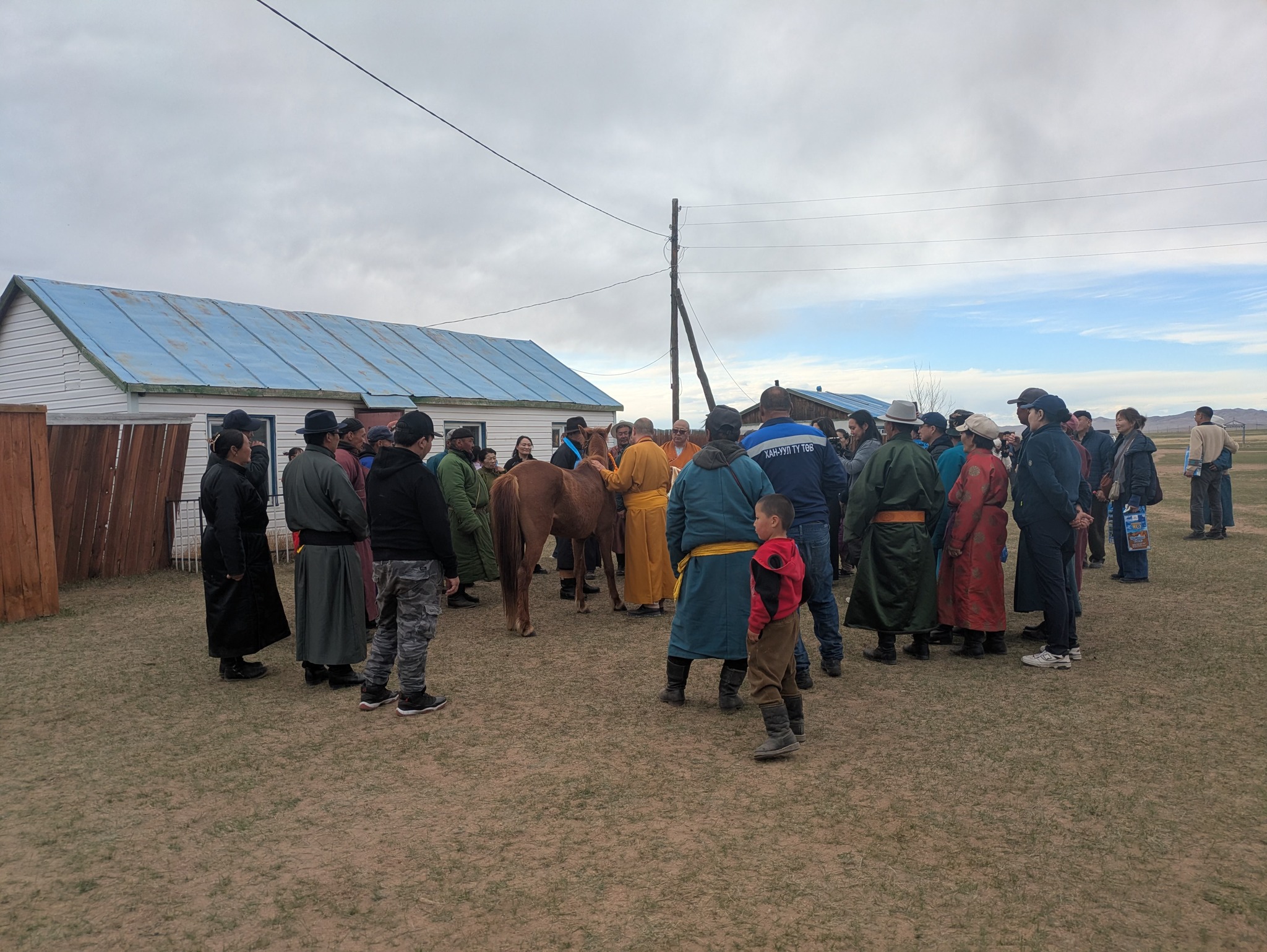 A photo of people of all ages in a Mongolian village greeting a Buddhist monk wearing saffron yellow robes.
