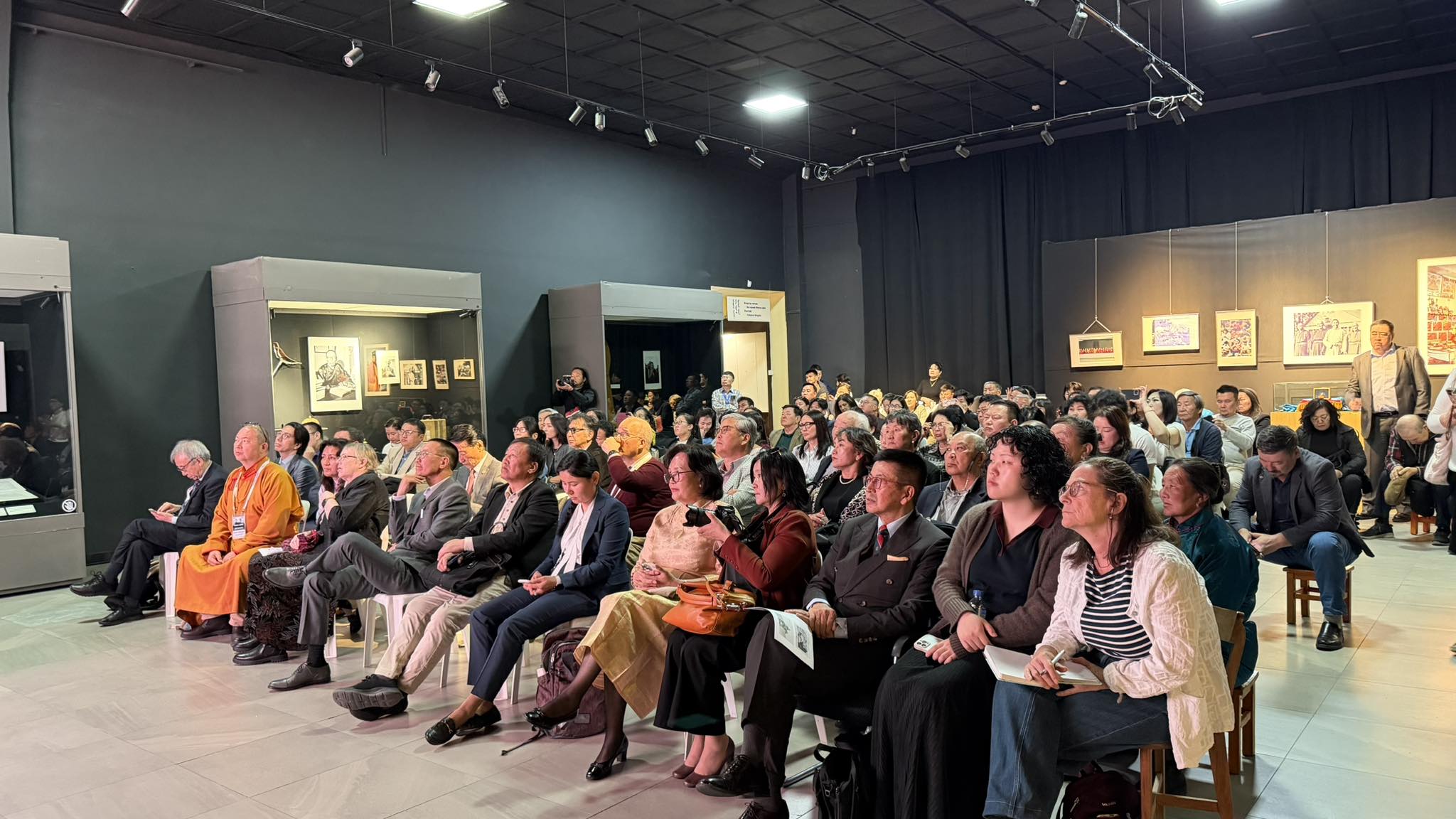 A crowd of people sitting inside a museum exhibition hall listening to a presentation.