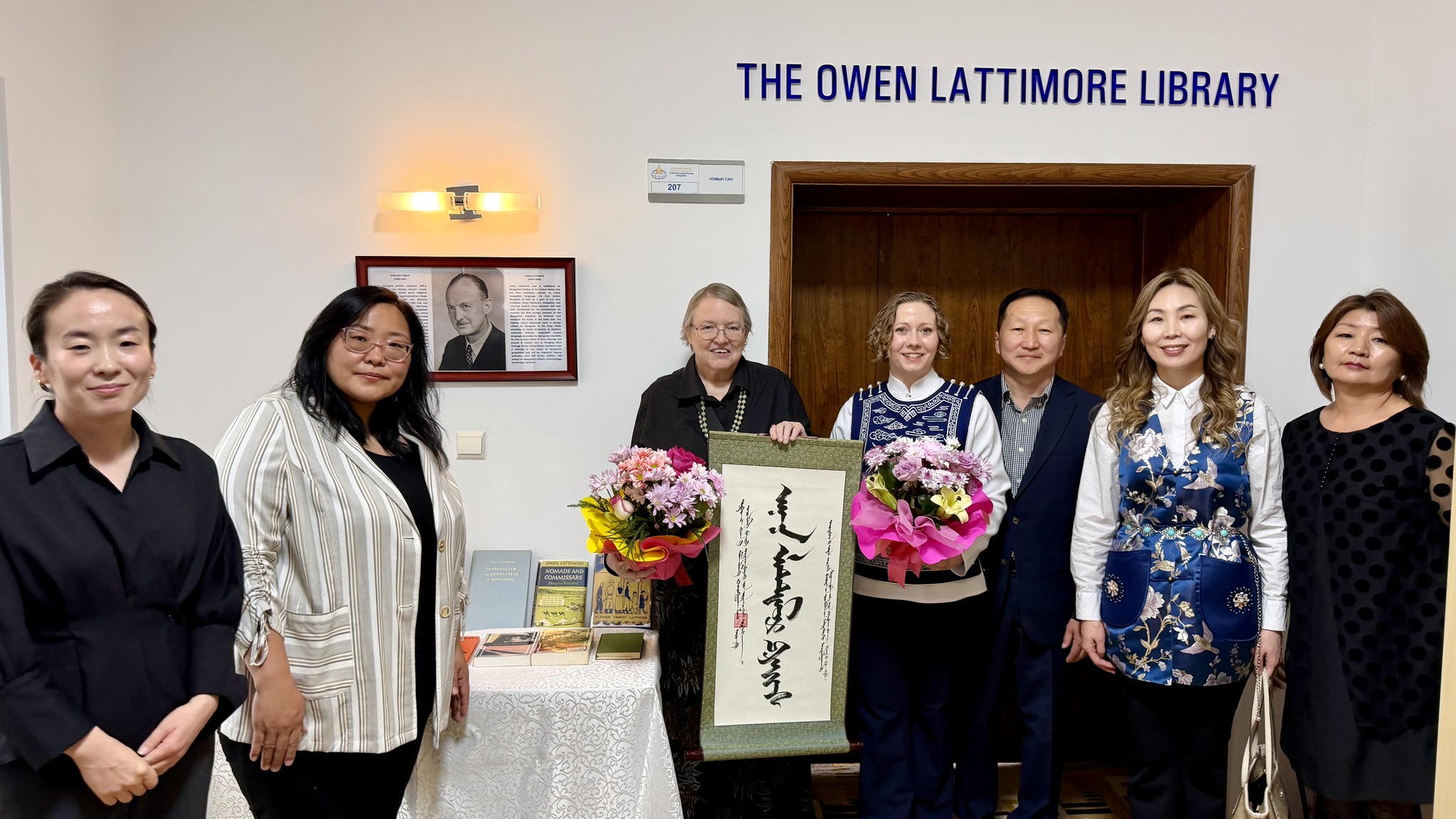 A group of six women and one man stand in a room with the sign “The Owen Lattimore Library.“ An older American woman holds flowers and a scroll and poses next to a table of books.