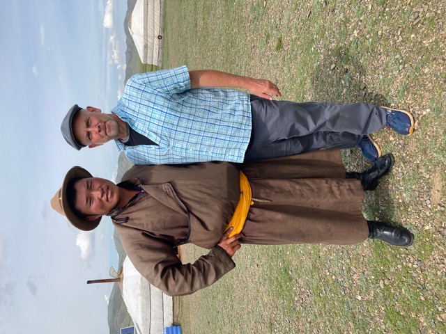 A Mongolian man in a brown robe and a Caucasian man in a blue shirt and gray trousers stand outside in front of traditional Mongolian gers posing together for the camera