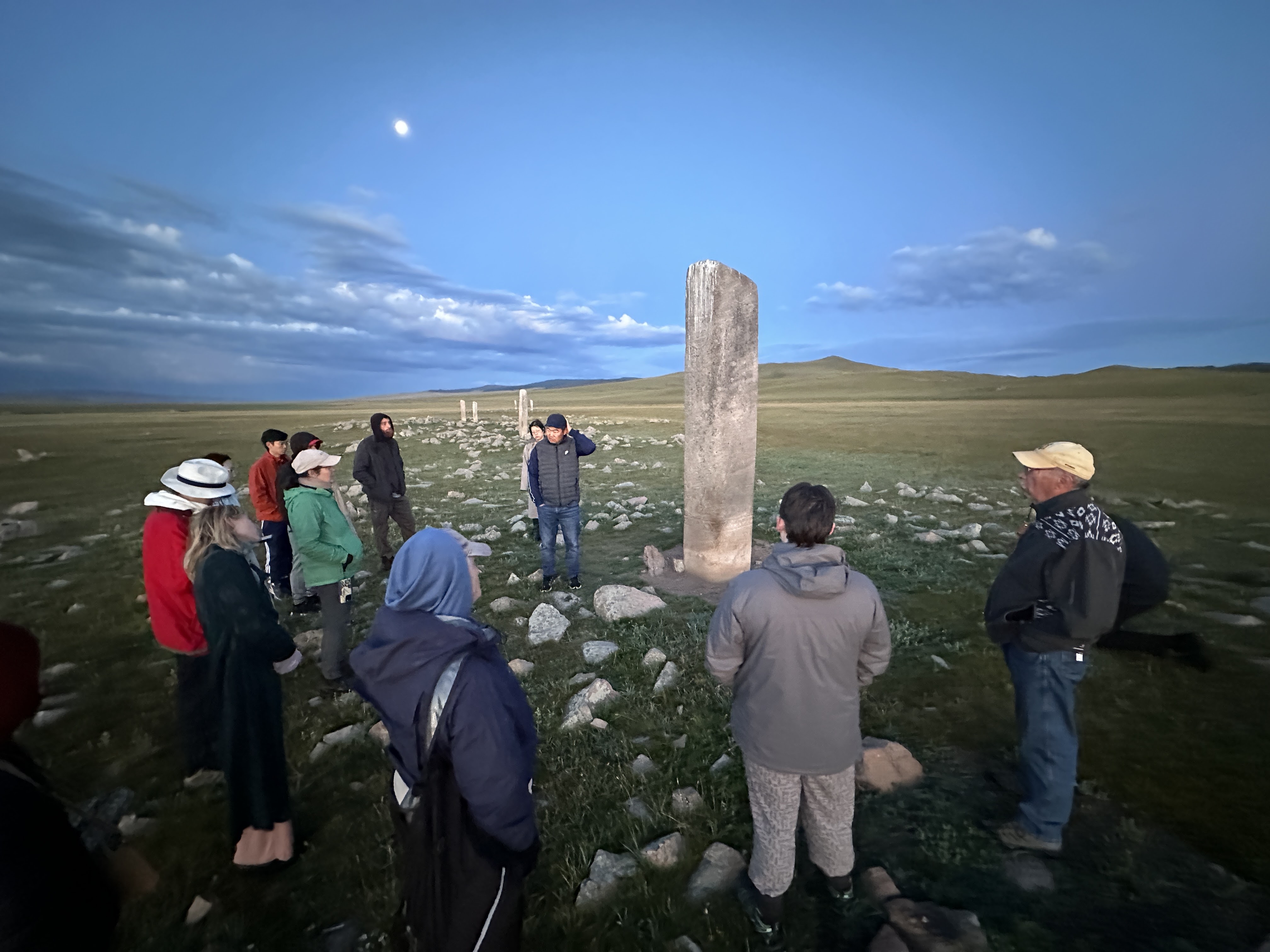 A group of people are standing outside looking at a tall, ancient, gray stone monument.