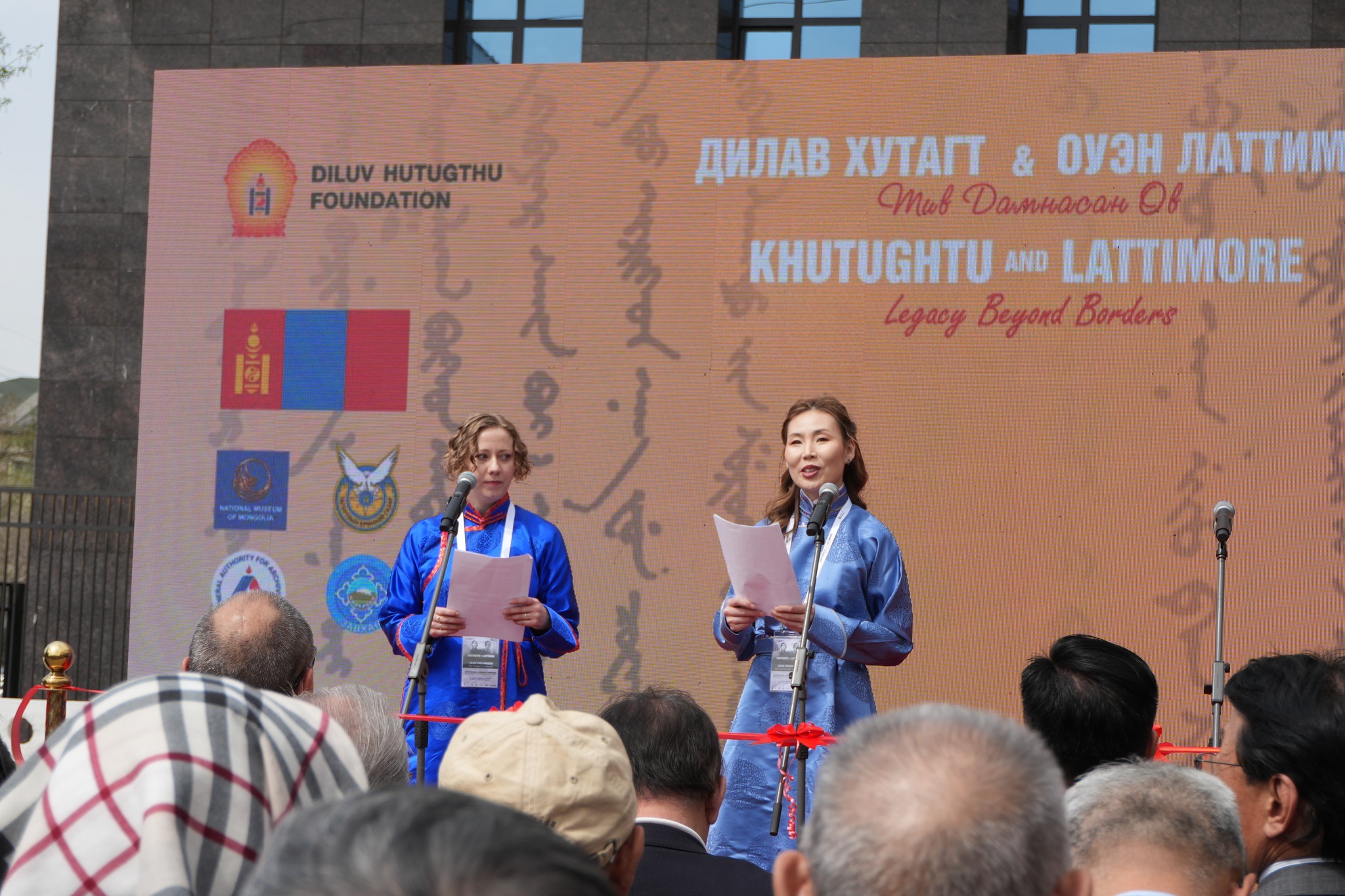 A photo of two female-presenting people standing onstage in traditional Mongolian clothing speaking in front of a crowd of people.