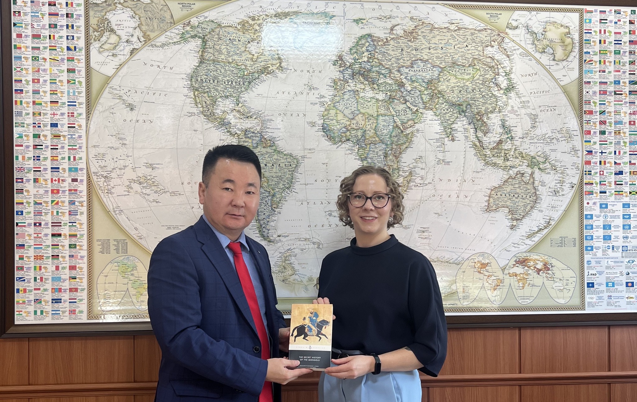 Professor Zayabaatar Dalai (left) and Maggie Lindrooth (right) stand in front of a world map holding up a copy of “The Secret History of the Mongols“ together.