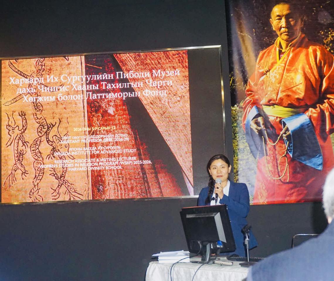 A young Mongolian woman standing behind a desk holding a microphone and giving a presentation.