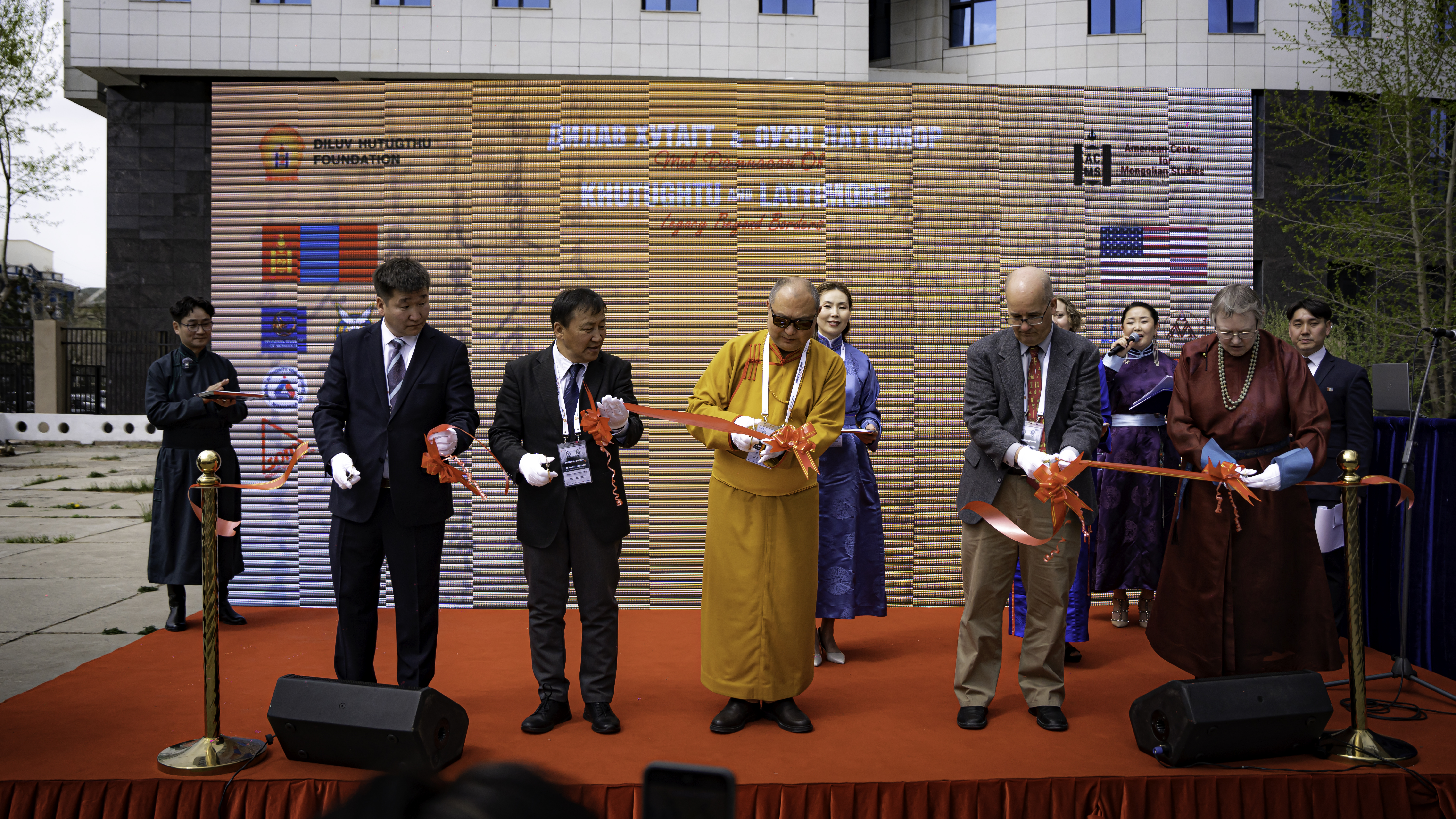 A group of people standing on an outdoor stage cutting a red ribbon with scissors.