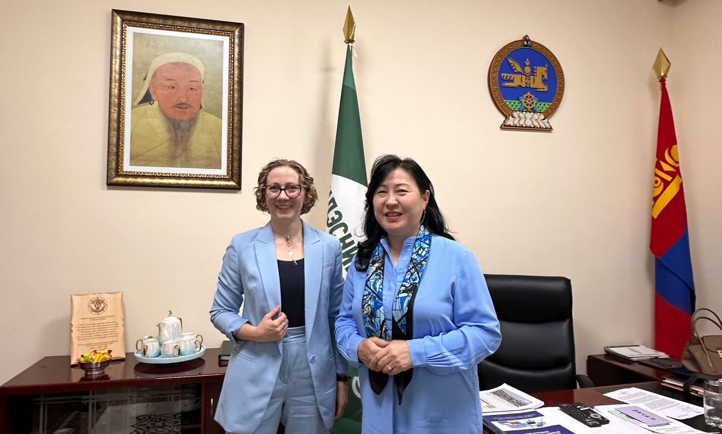 Maggie Lindrooth (left) and L. Undraa (right) stand in front of a portrait of Chinggis Khan and the emblem of Mongolia