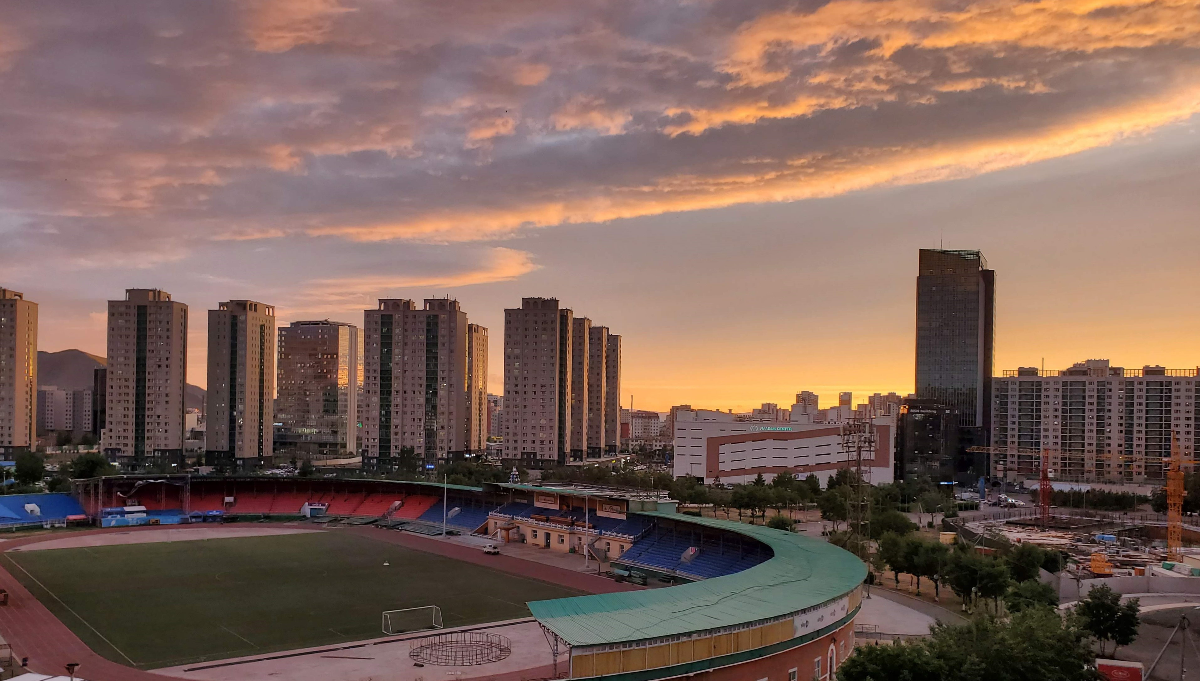 A view of Ulaanbaatar city's skyline at sunset. Tall buildings stand in the background and a sports stadium is in the foreground.