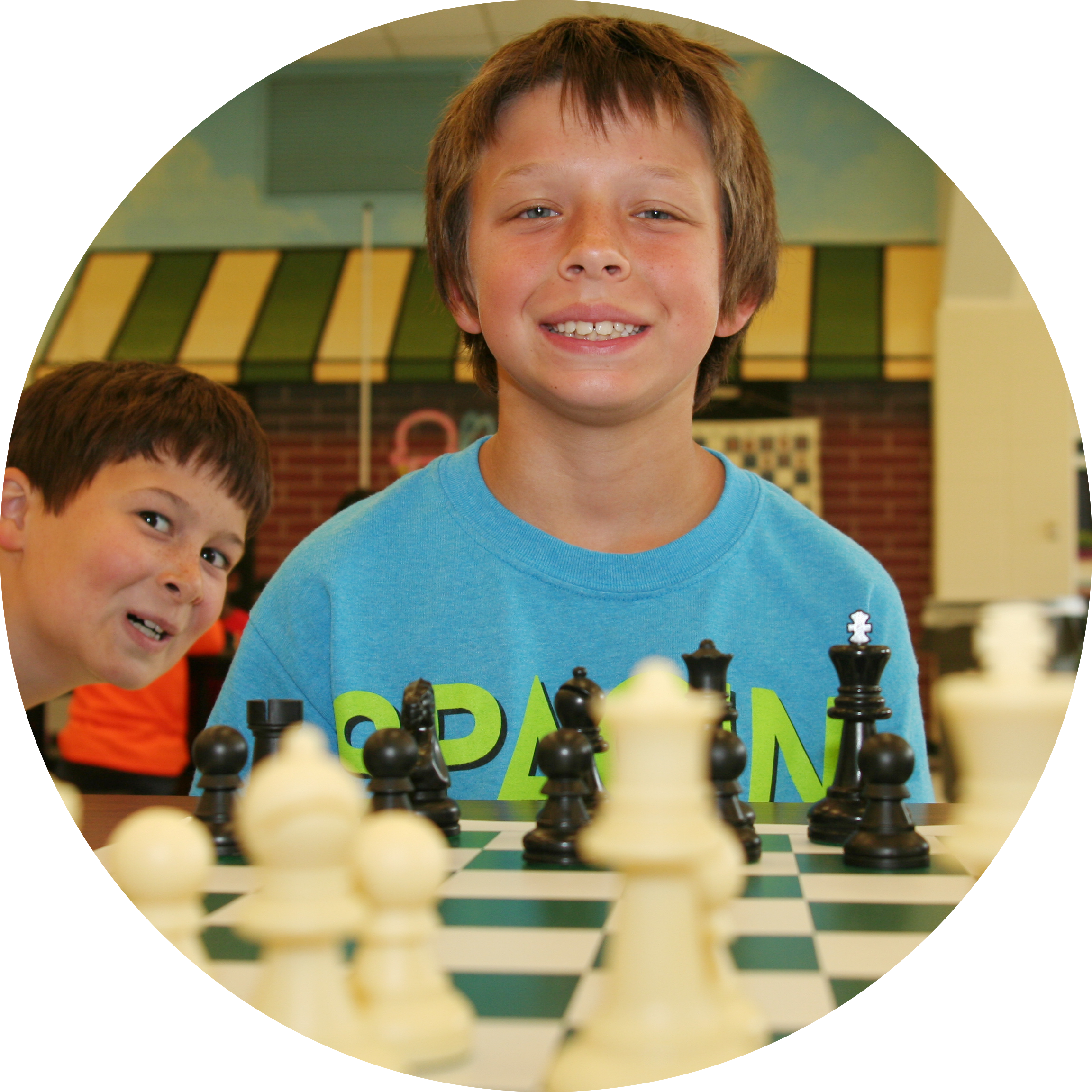 Boy in a blue shirt grinning over a chess board with a buddy photobombing from the left.