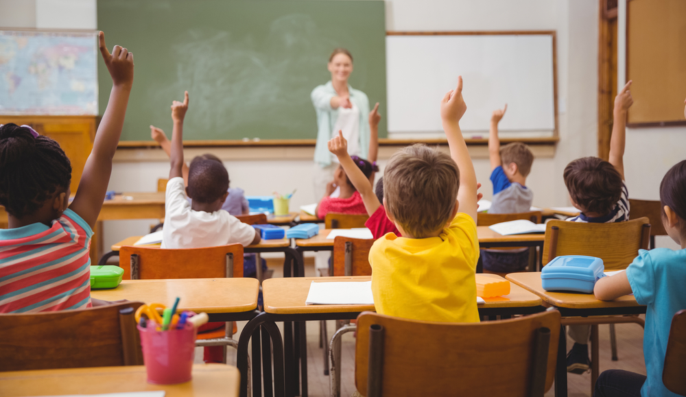 Children in a classroom raising hands.  Teacher is standing at the front of the room by the chalkboard.