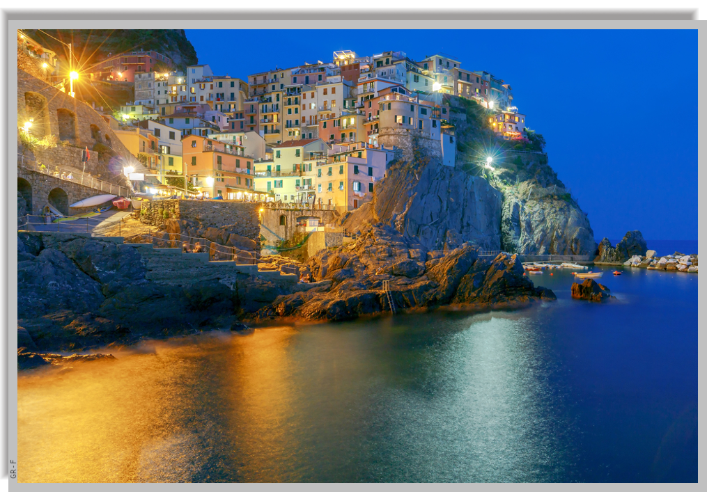 The village of Manarola, perched on a rocky hill overlooking the sea, with its colorful houses nestled together. Nighttime shots with lights reflecting on the calm sea.