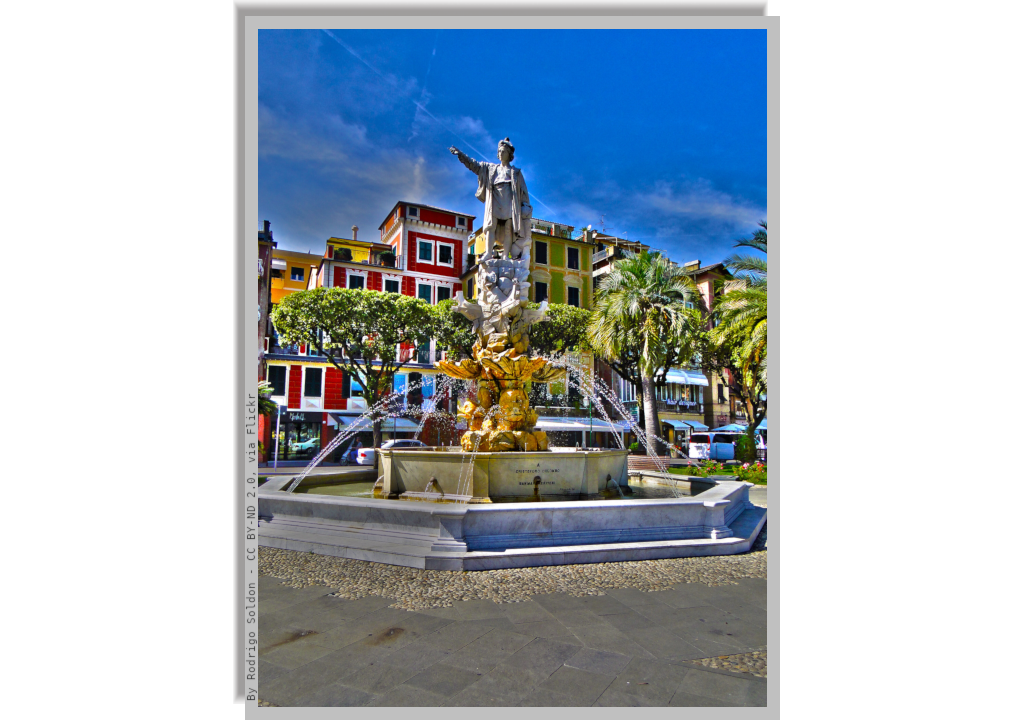 Monument to Christopher Columbus in a square with fountain, maritime pine trees and houses in the background