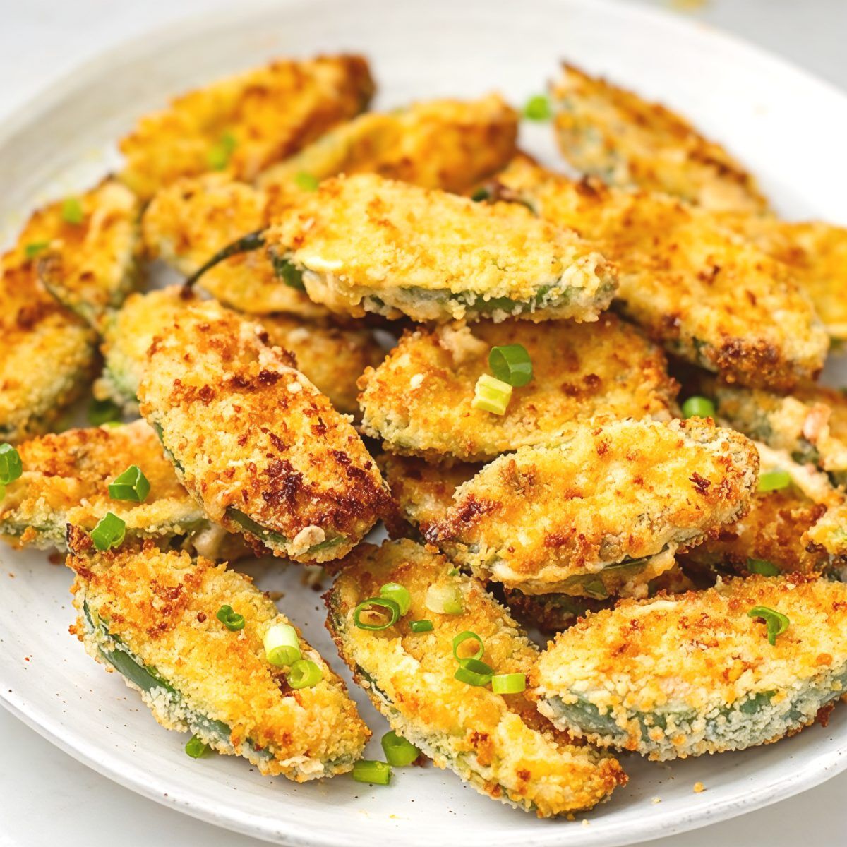 Crispy breaded jalapeño poppers stuffed with melted cheese and sprinkled with chopped green onions on a white serving plate, making a delicious party appetizer.