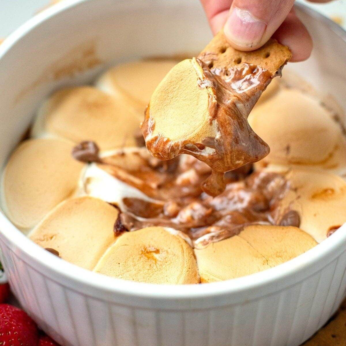 A hand dips a graham cracker into a white ramekin of warm s'mores dip, scooping up melted chocolate and toasted marshmallows.