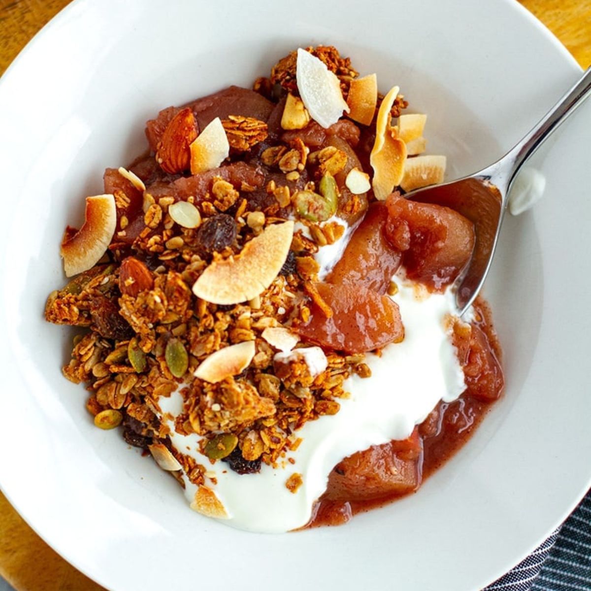 Bowl of warm spiced stewed apples topped with crunchy granola, coconut flakes, and a dollop of yogurt being scooped with a spoon.