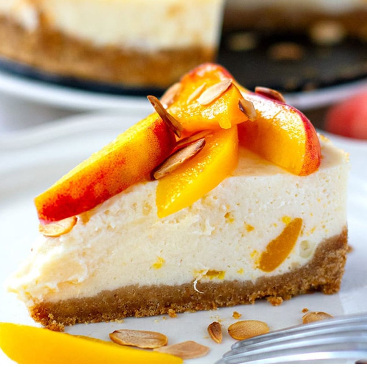 Slice of creamy peach cheesecake on a white plate topped with fresh peach slices and toasted almonds with a graham cracker crust.