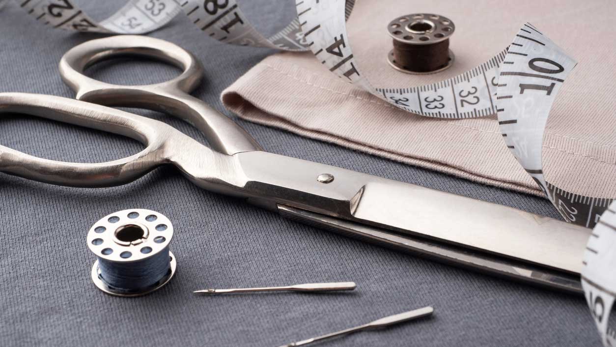 A stainless steel pair of scissors sitting on gray knit fabric with a bobbin, a curly measuring tape and a few sewing machine needles deoratively placed