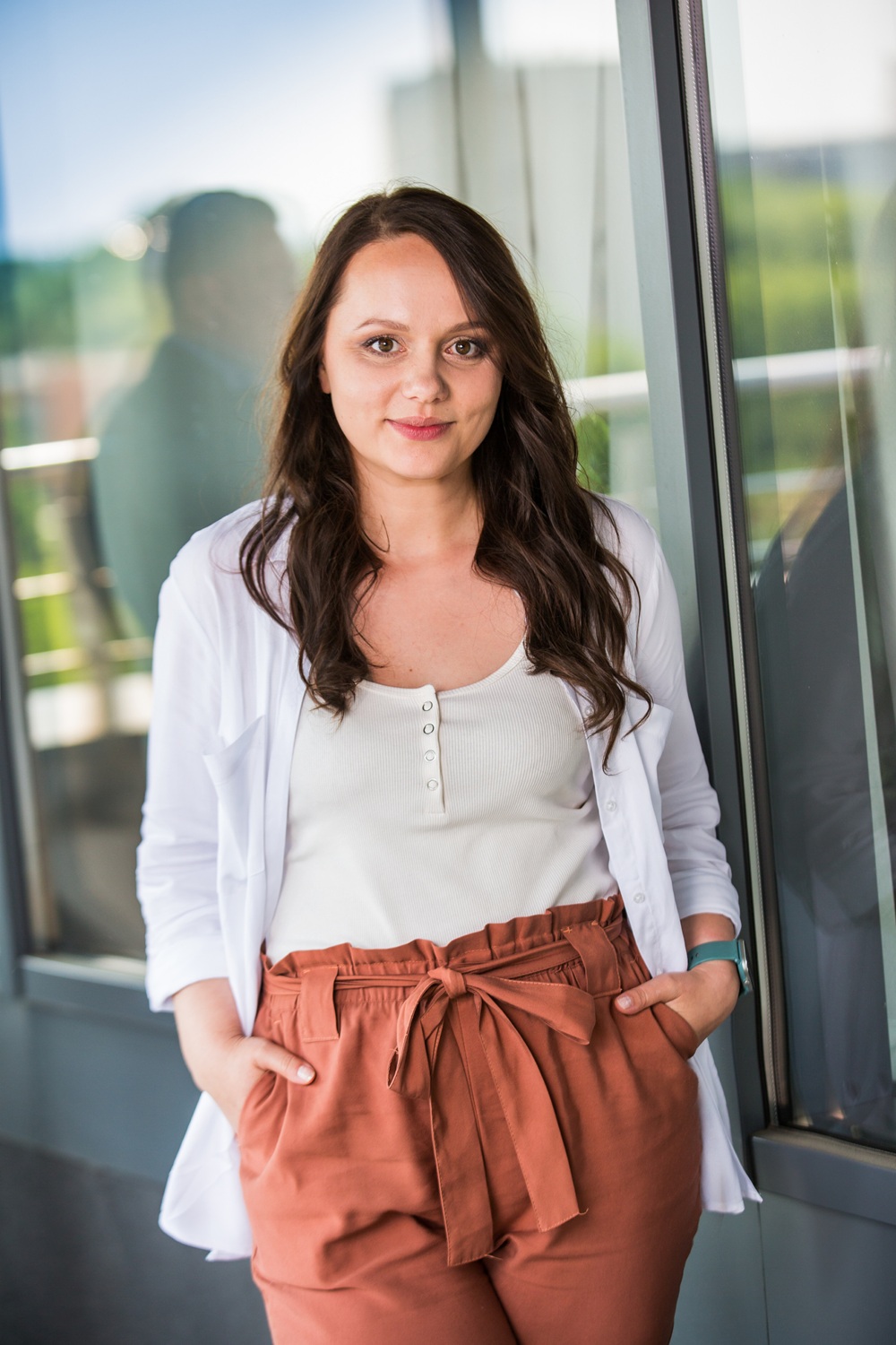 Uśmiechnięta kobieta z długimi, ciemnymi włosami pozuje na tle nowoczesnego budynku. Ma na sobie elegancką, białą koszulę z krótkim rękawem i beżowe spodnie. Stoi z założonymi rękami, patrząc pewnie w stronę kamery.