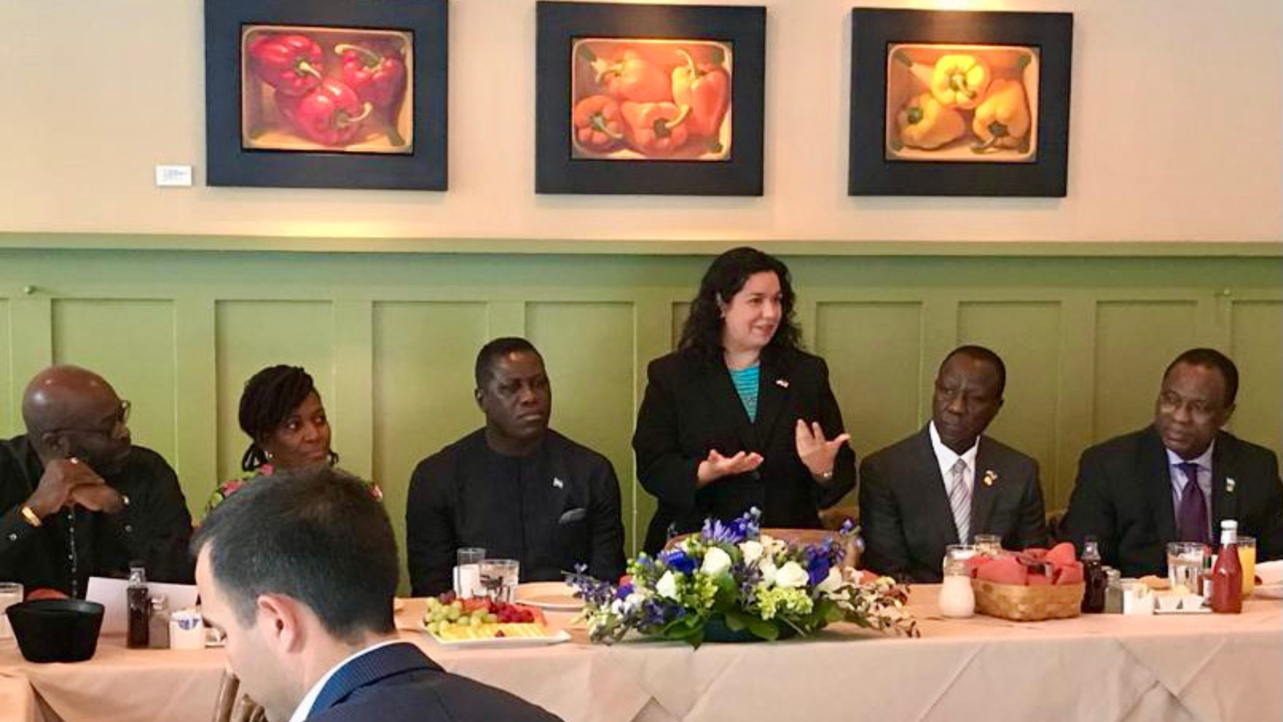 A woman in a blazer stands speaking at a meeting, with five people seated around a table set with fruit and drinks, as framed bell pepper art hangs on the wall behind them.