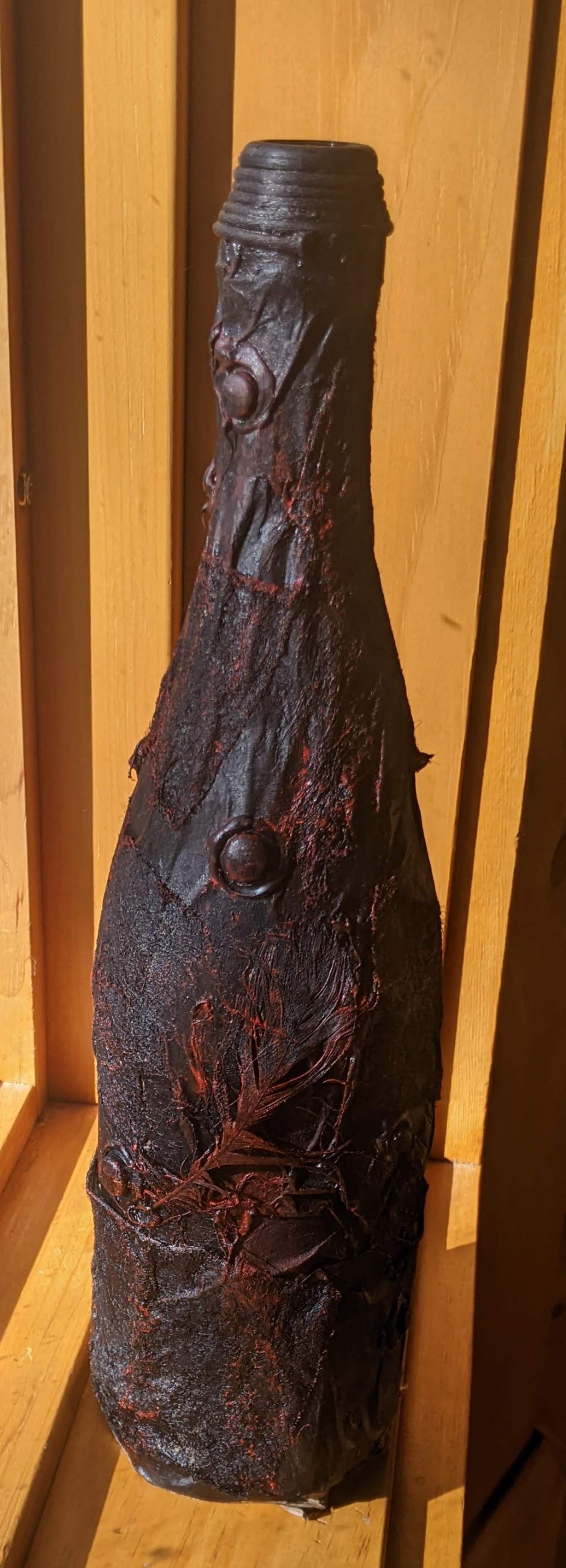 An old sparkling grape juice bottle covered in brown packing paper, googly eyes, dryer sheets, some glue, and a feather, all painted black, then splattered with red paint.