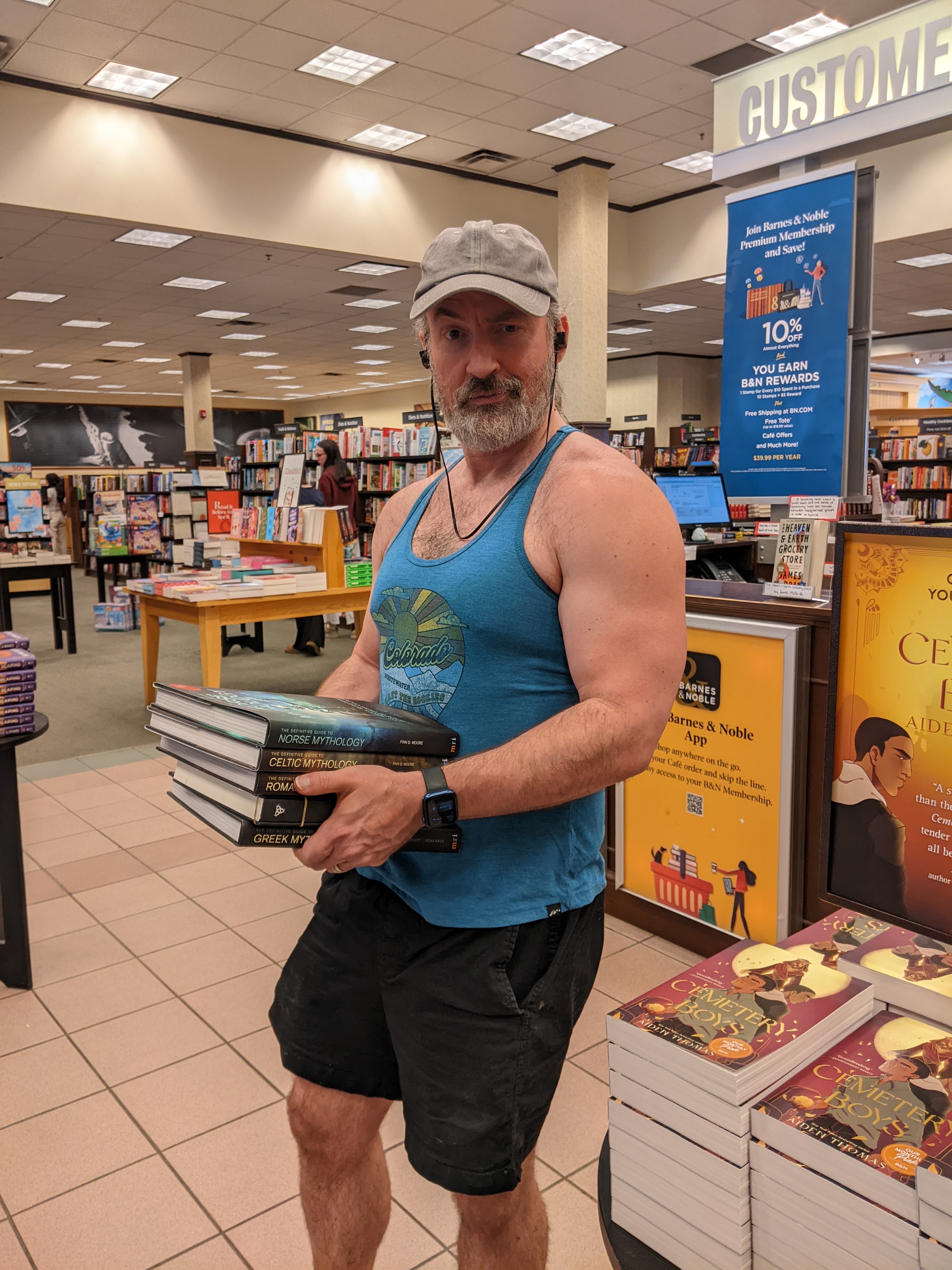 Picture of Jim standing in Barnes and Noble in a tank top and massive biceps holding a stack of mythology books.