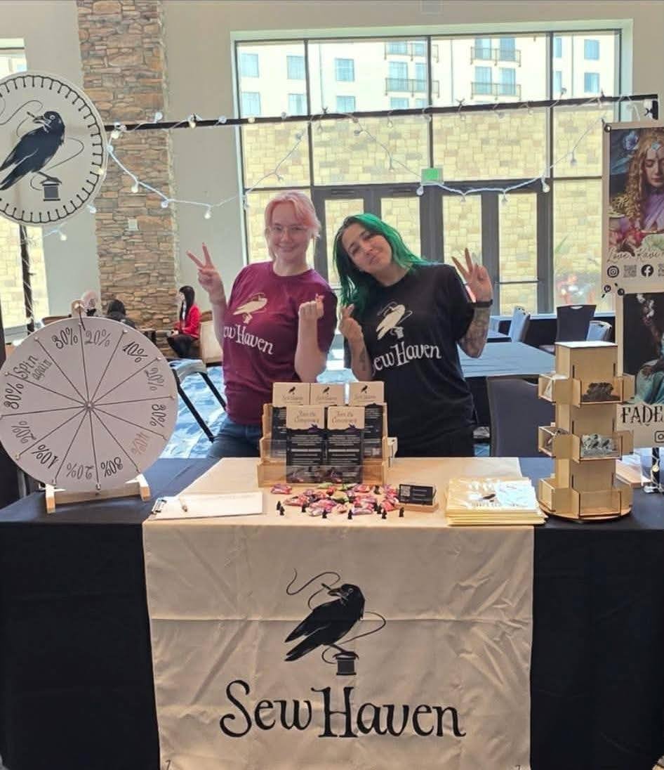 Kavi and a friend standing at a Sew Haven table at a local convention, looking like happy nerds.