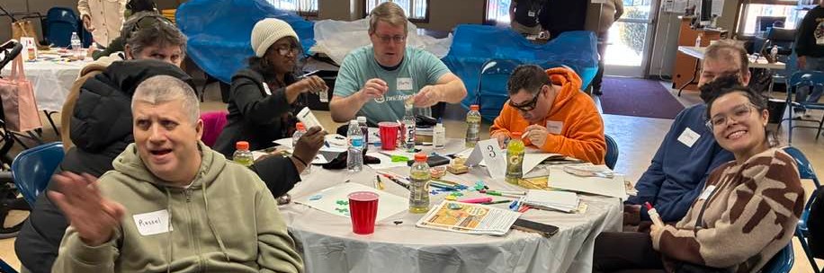 A group of people around a table working on projects some smiling and waving at the camera.