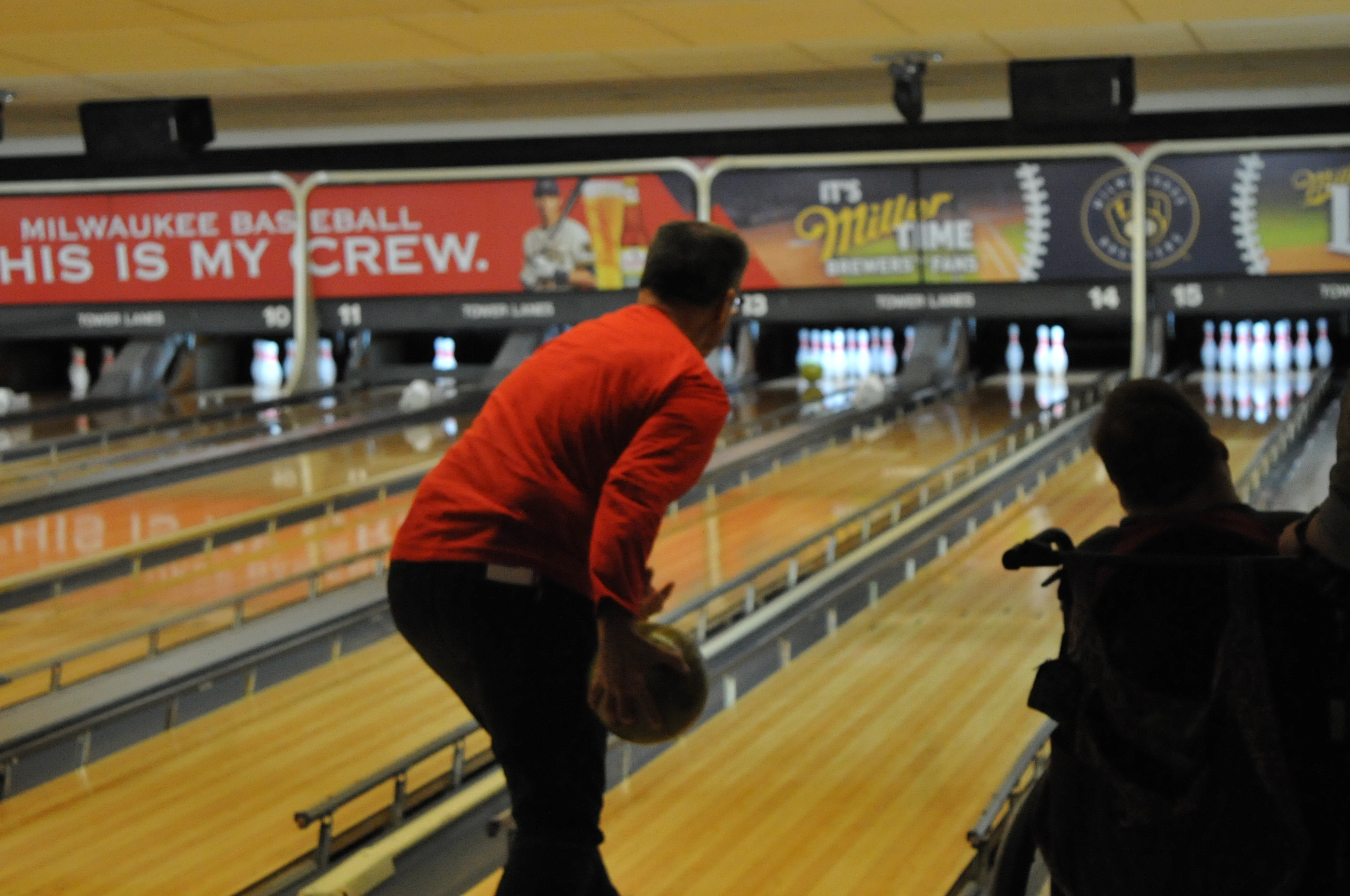 Looking from behind at a man about to throw a ball while a person in a wheelchair on the bowling lane right next to them