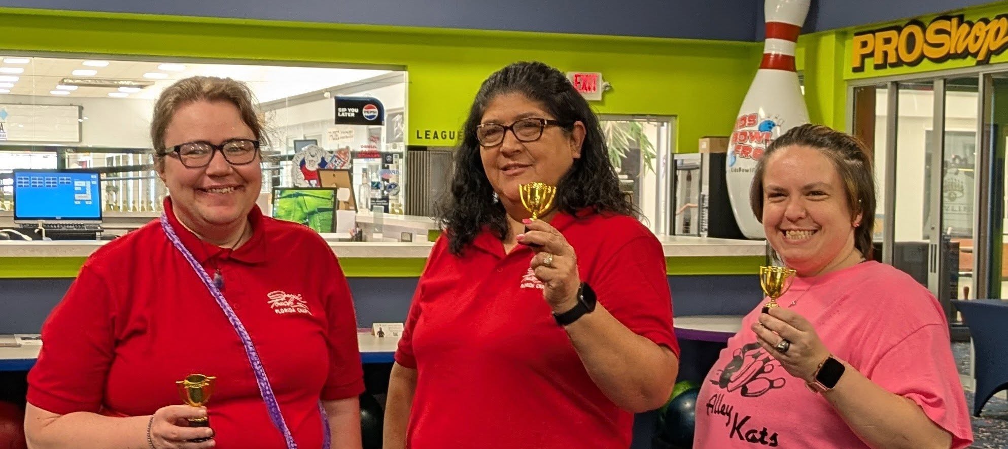three women at a bowling alley holding up tiny trophies