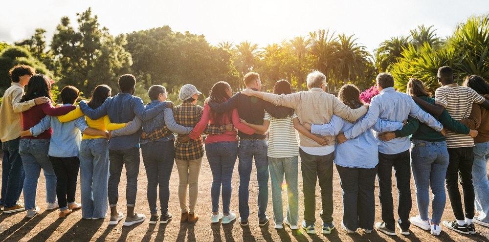 A group of people of all generations and abilities hugging each other in a half circle outdoors in the bright sunshine