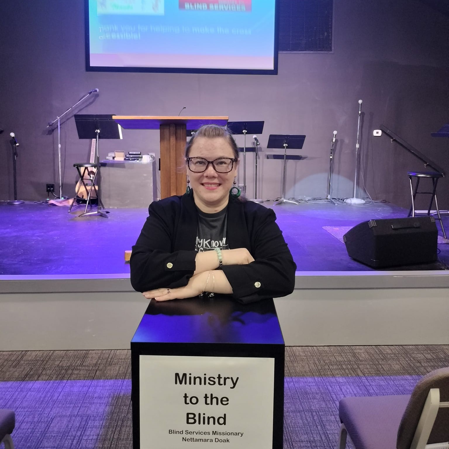 Nettamara in front of a church with a podium that reads Ministry to the Blind Blind Services Missionary Nettamara Doak